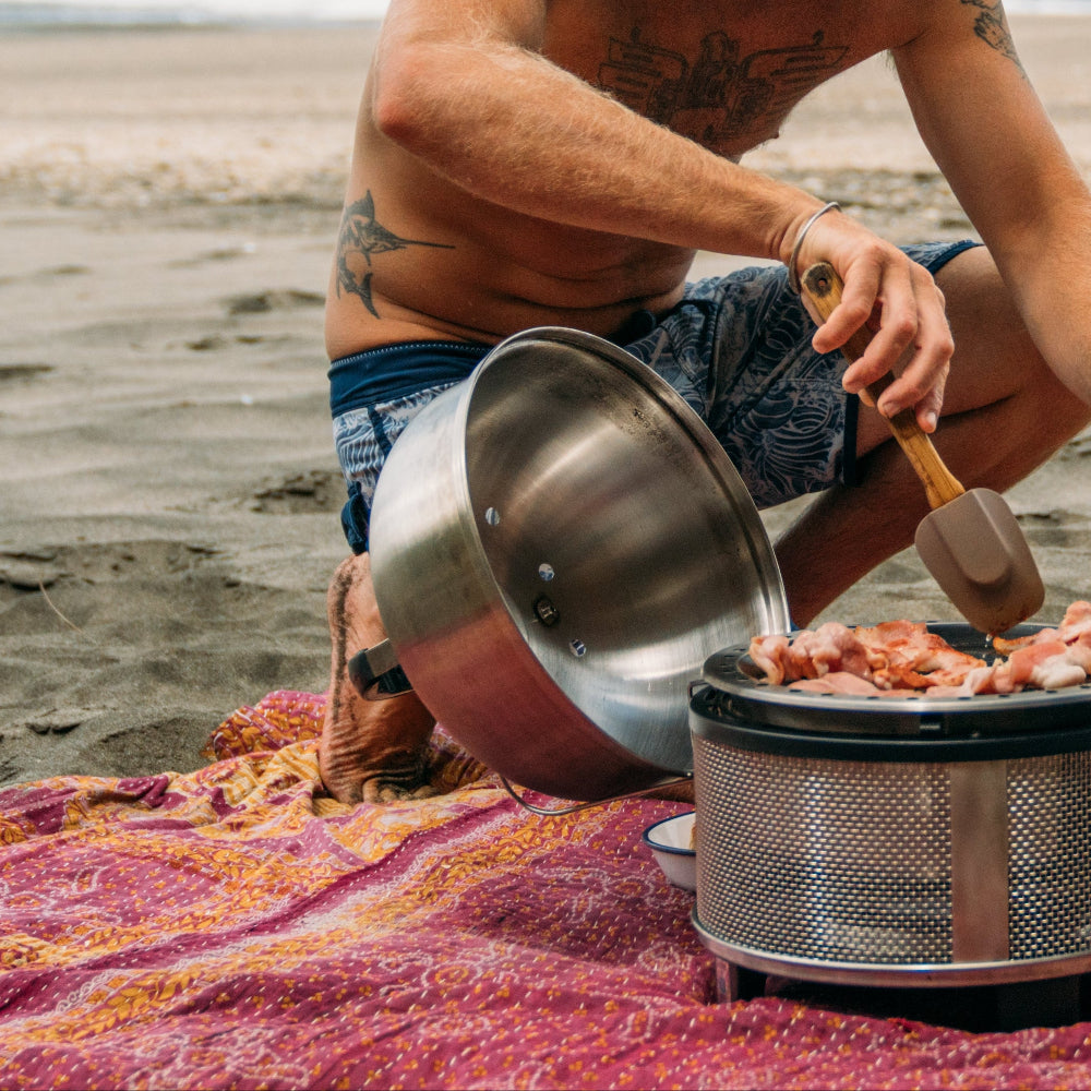 Man cooking outdoors on a beach with a portable stove and pots.