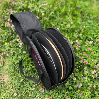 A black fabric carrying case holding several round black discs, photographed outdoors on grass with small pink flowers. The case has a visible red-and-white logo that reads COBB.