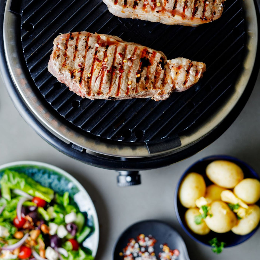 Grilled meat on a COBB barbecue grill with side dishes including salad and potatoes.