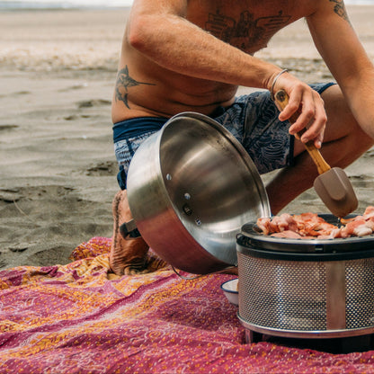Man cooking outdoors on a beach with a portable stove and pots.