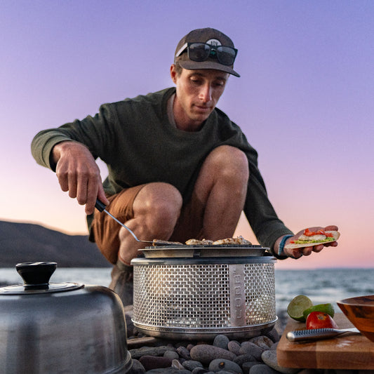Person cooking using a COBB outdoors on a rocky beach with a sunset in the background