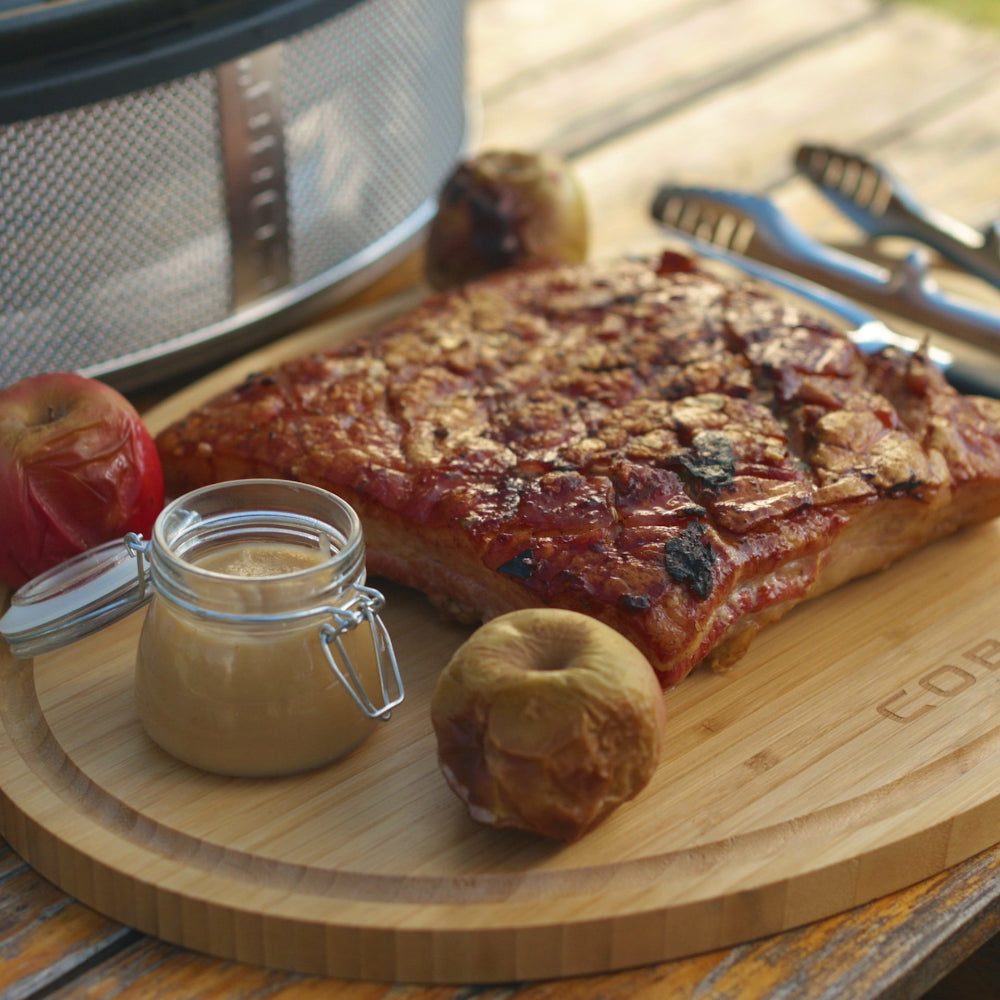 Grilled meat on a wooden cutting board with apples and a jar of sauce, outdoors.