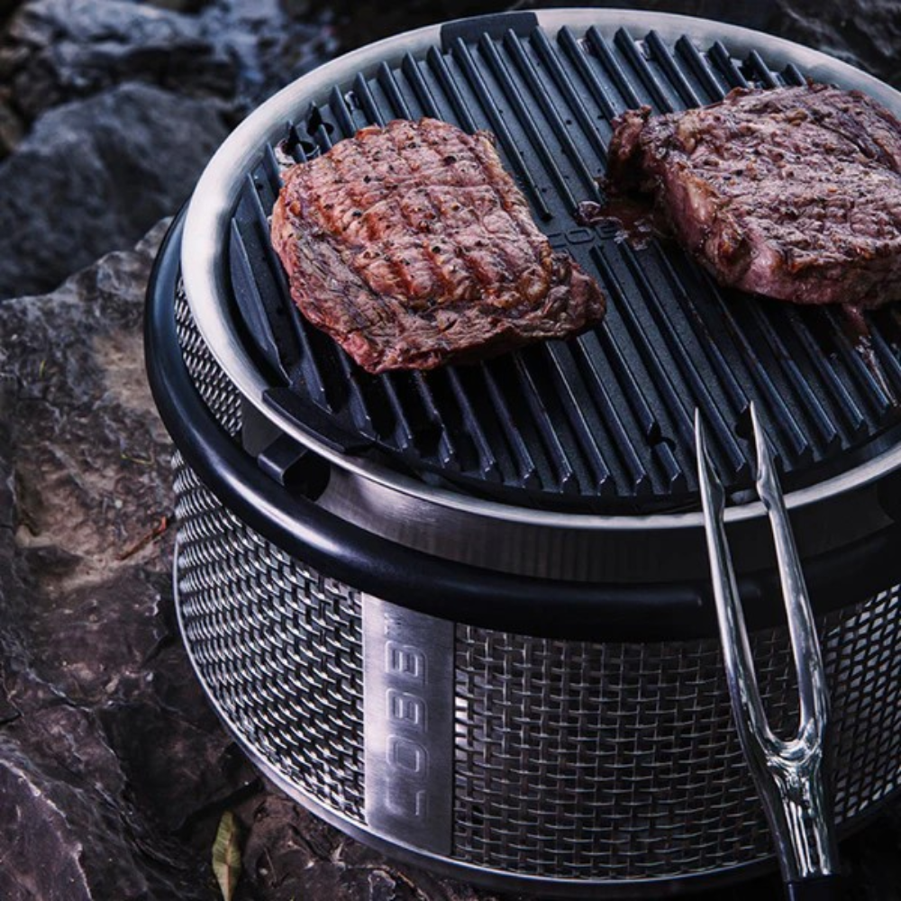Portable COBB grill with steaks cooking on a rocky surface